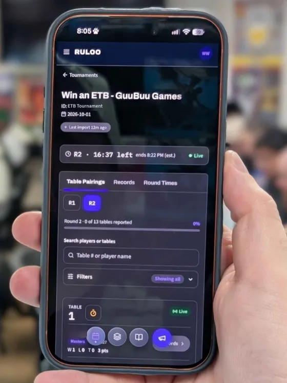A hand holding a phone showing Ruloo's live table pairings and round clock at a Pokémon TCG event.